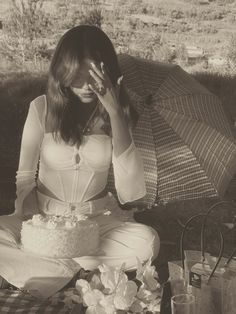 a woman sitting on the ground with an umbrella over her head and cake in front of her