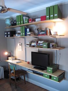 a room with a desk, bookshelf and computer monitor on top of it