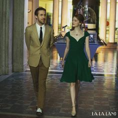 a woman in a green dress and a man in a beige suit walking down a hall