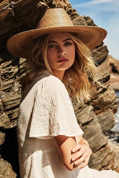 a woman wearing a hat and sitting on rocks near the ocean with her hands in her pockets