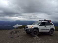 a white suv parked on top of a mountain under a cloudy sky with mountains in the background