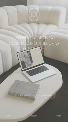 an open laptop computer sitting on top of a white table next to a book and couch