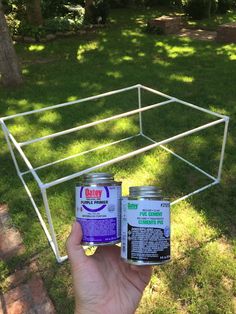 a hand holding two cans of paint in front of a white metal rack on the grass