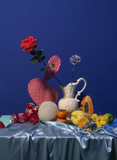 an arrangement of flowers and fruit on a blue table cloth with a heart - shaped vase