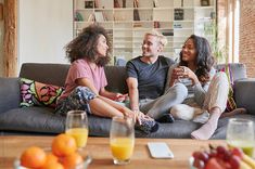 three people sitting on a couch with fruit and drinks in front of them, smiling at each other