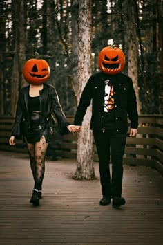 two people wearing pumpkin heads walking on a bridge in the woods with their hands held by each other