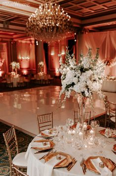 a wedding reception setup with white flowers and gold napkins on the table in front of a chandelier