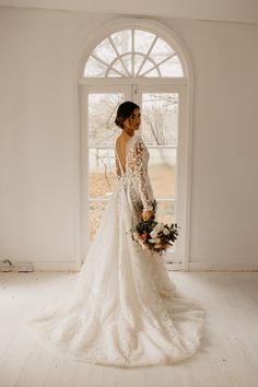 a woman standing in front of a window wearing a wedding dress and holding a bouquet