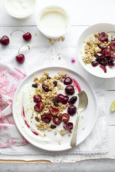 yogurt, granola and cherries are served on a white plate with spoons