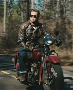 a man riding on the back of a red motorcycle down a road next to trees