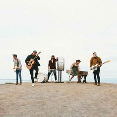 a group of people standing on top of a sandy beach next to the ocean playing guitar