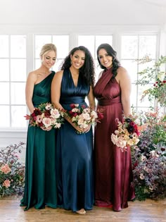 three women in long dresses standing next to each other with flowers and greenery behind them