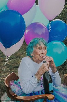 an older woman sitting in a chair holding a bottle of wine with balloons behind her