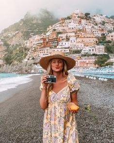 a woman in a dress and hat taking a photo on the beach with an old camera