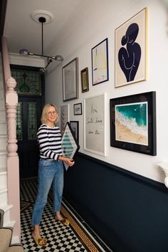 a woman standing next to a wall with pictures on it and holding a framed photo