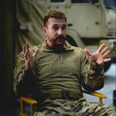 a man sitting in a chair with his hands out and an army truck behind him