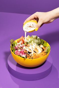 a person pouring dressing into a salad in a yellow bowl on a purple tablecloth