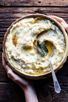 two hands holding a bowl of mashed potatoes with sage leaves on the side and a spoon in it