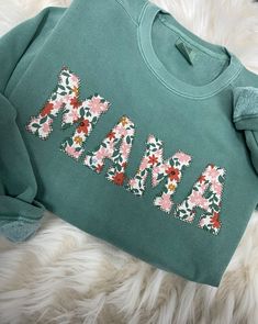 a green sweatshirt with flowers and the word mama on it is laying on a furry surface