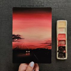 someone is holding up a card with the sunset in the background and some watercolor paints on it