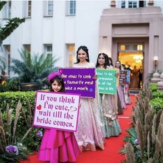 several women in dresses holding signs on a red carpeted walkway with plants and bushes