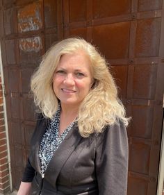 a woman with blonde hair standing in front of a brick wall wearing a black jacket