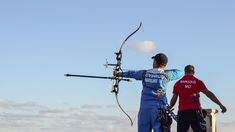 two men standing next to each other while holding an arrow and aiming something in the air