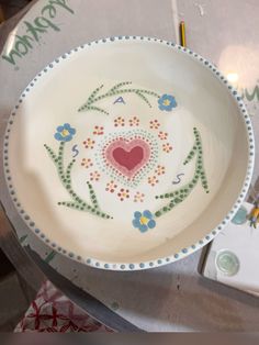 a white bowl with a heart painted on the side and flowers in the middle is sitting on a table