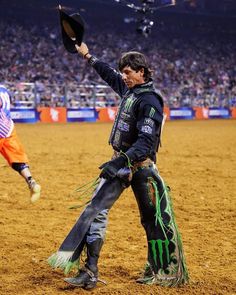 a man in black jacket holding up a hat on top of a dirt covered field