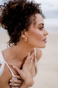 a woman with curly hair and earrings on her neck looking off into the distance at the beach