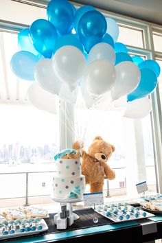 a teddy bear sitting on top of a table with blue and white balloons in the air