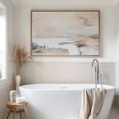 a white bath tub sitting next to a window in a bathroom with a painting on the wall