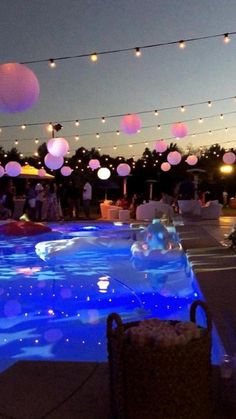 people are gathered around an illuminated swimming pool at night with paper lanterns hanging above it