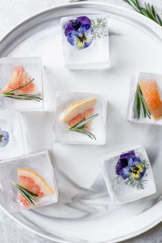 four square ice cubes with flowers and herbs in them on a white plate surrounded by greenery