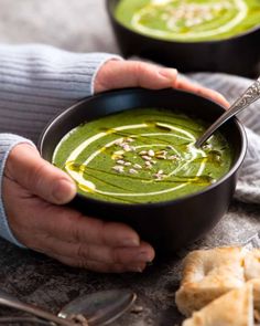 a person holding a bowl of green soup with pesto and almonds on the side