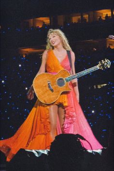 a woman in an orange and pink dress holding a guitar while standing on top of a stage