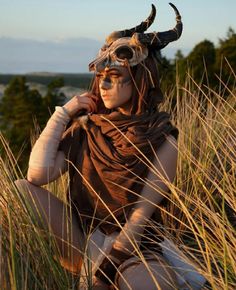 a woman with horns is sitting in tall grass wearing a mask and scarf on her head