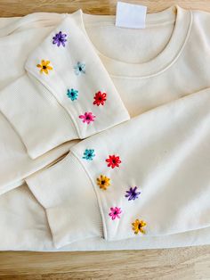 two white sweaters with colorful flowers on them sitting on a wooden floor next to each other