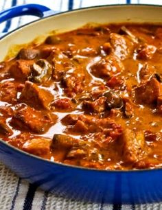 a blue pan filled with meat and sauce on top of a tablecloth next to a spoon