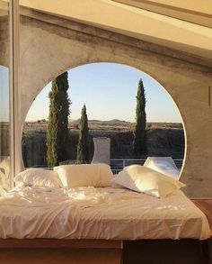 a bed with white sheets and pillows sitting in front of a large round window that looks out onto the countryside