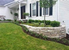 a stone wall in front of a white house with green grass and bushes around it