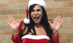 a woman wearing a santa hat and holding her hands in the air while standing next to a brick wall