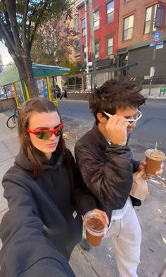 two people taking a selfie while holding drinks and talking on their cell phones in the street