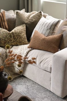 a white couch with lots of pillows on it's back and some plants in the corner