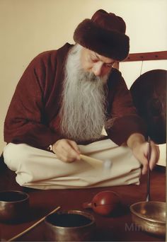 an old man with a long white beard sitting at a table and writing on paper