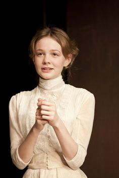 a woman in a white dress standing with her hands folded and looking at the camera