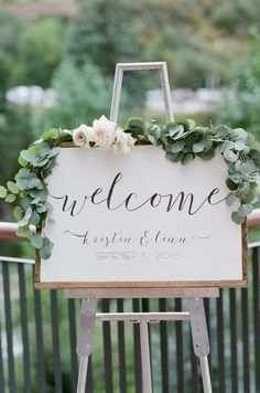 a welcome sign with greenery and flowers is displayed on a wooden easel for an outdoor ceremony