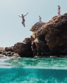 two people jumping off rocks into the ocean with their arms in the air while another person is