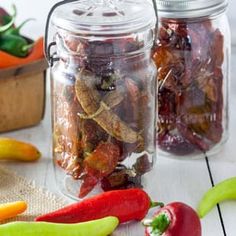 three glass jars filled with different types of peppers and peppers on a white wooden table