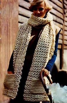 a woman wearing a crocheted scarf and holding a cane in her hand while standing next to a wooden fence